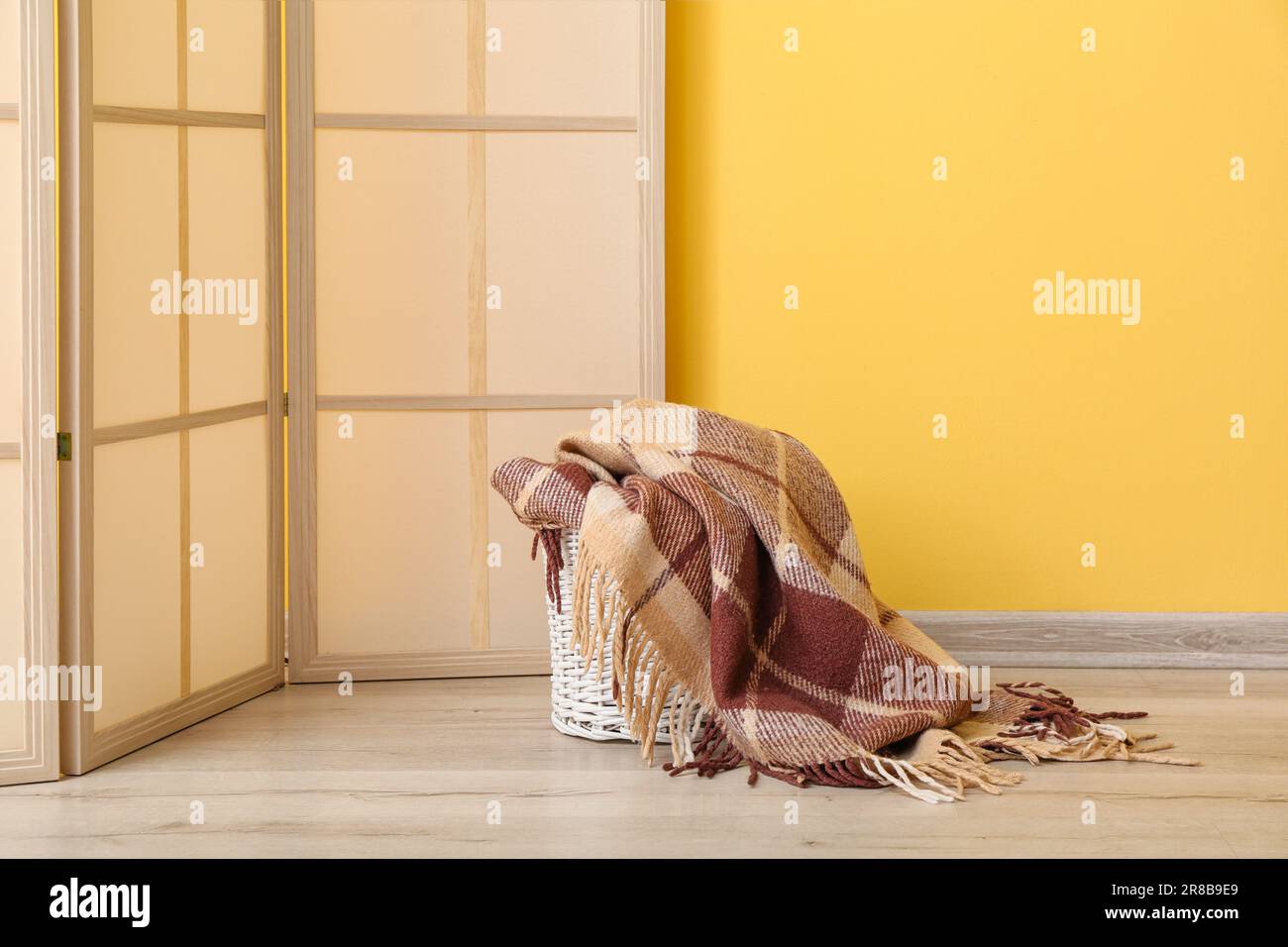 Cestino con nuova coperta morbida e schermo pieghevole vicino alla parete gialla in camera Foto Stock