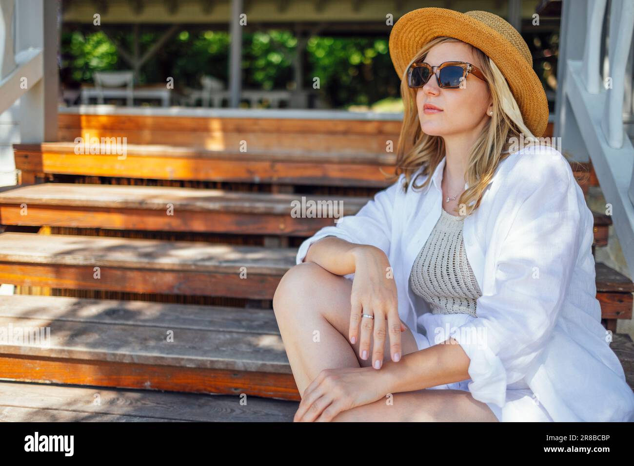 Donna bella fiduciosa con cappello di paglia e vestiti leggeri che guardano campi con cespugli di uva. Bella ragazza bionda in occhiali da sole si appoggia su rai in legno Foto Stock