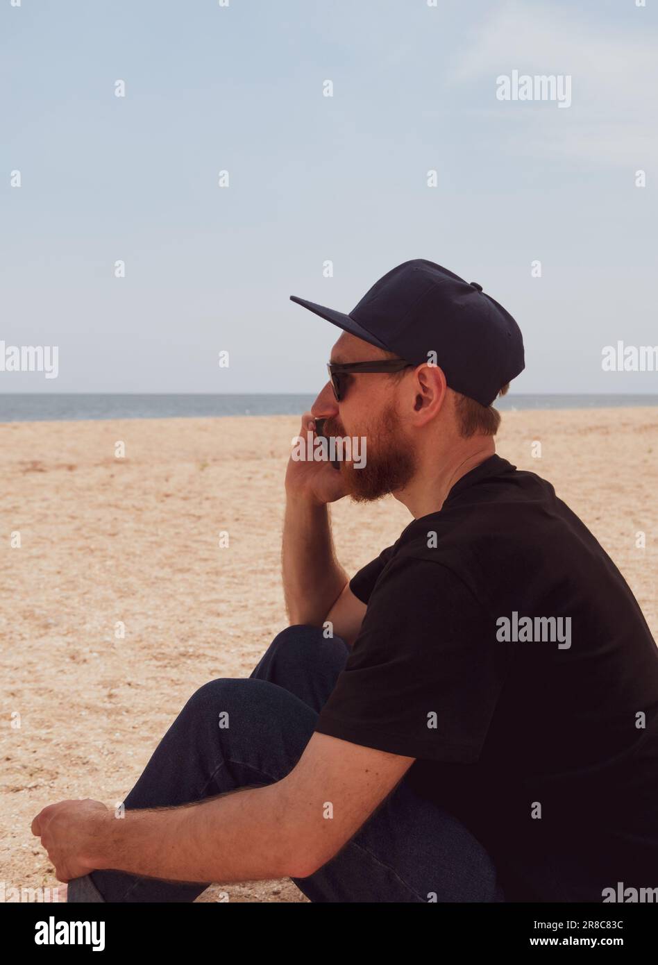 Uomo millenario beared in t-shirt nera snapback sul mare blu paesaggio con smartphone in mano parlando. Giovane adulto che chiama amici sul cellulare, Foto Stock