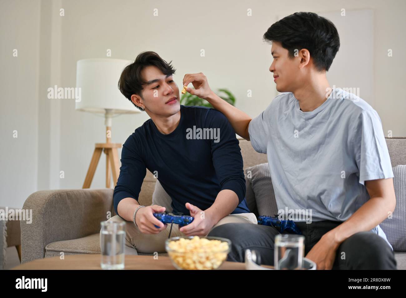 Un giovane gay asiatico felice e gioioso alimenta popcorn al suo ragazzo mentre si diverte nel soggiorno insieme. LGBTQ+, coppia gay, stile di vita Foto Stock