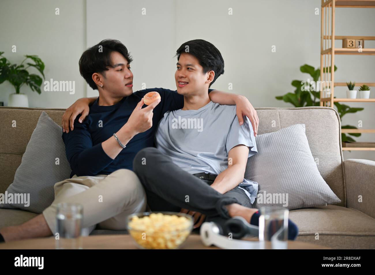 Un bel giovane gay asiatico, felice e simpatico, nutre una ciambella al suo ragazzo mentre si rilassa su un divano nel soggiorno insieme. LGBTQ+, stile di vita, c Foto Stock