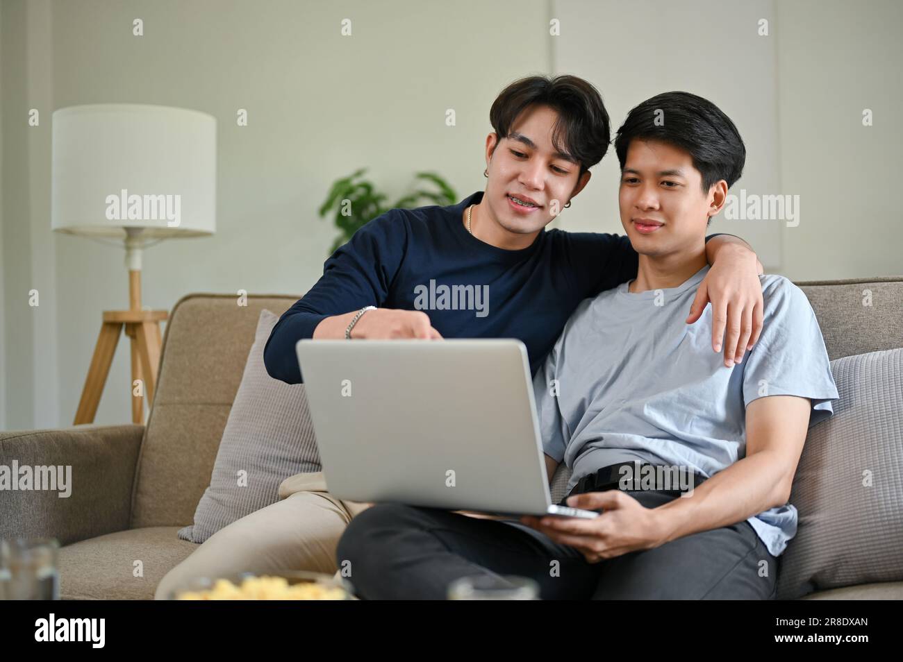 Un giovane gay asiatico felice ama guardare un film attraverso un computer portatile con il suo ragazzo su un divano nel soggiorno insieme. Foto Stock