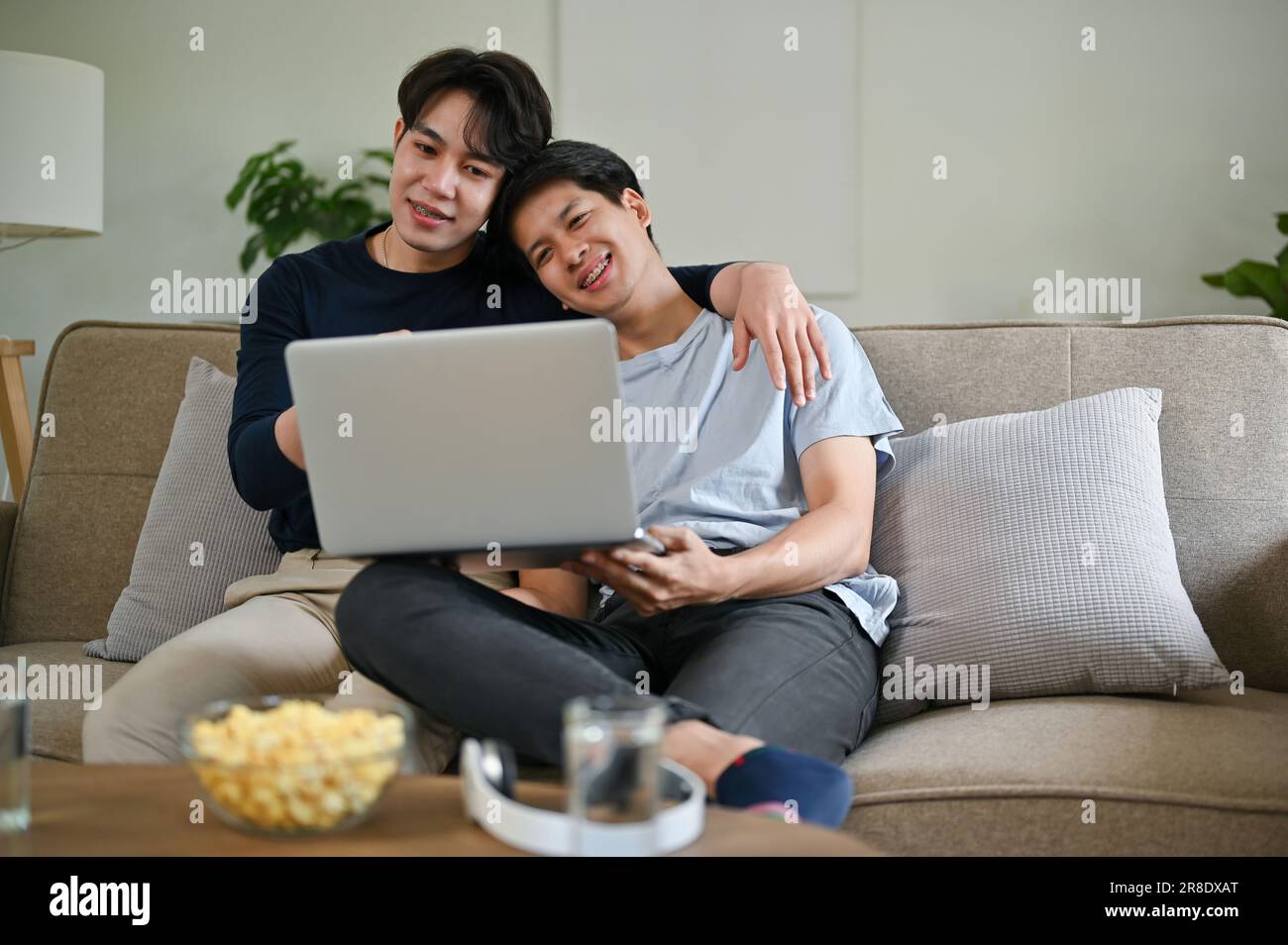 Un giovane gay asiatico felice si sta rilassando e abbracciando il suo ragazzo mentre si guarda un film attraverso un computer portatile su un divano nel toget soggiorno Foto Stock