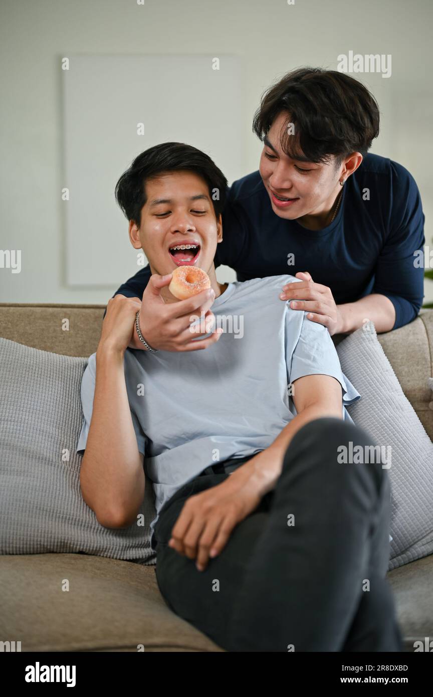 Un giovane gay asiatico felice alimenta una ciambella al suo ragazzo mentre si rilassa su un divano nel soggiorno insieme. LGBTQ+, lifestyle, coppia, relati Foto Stock