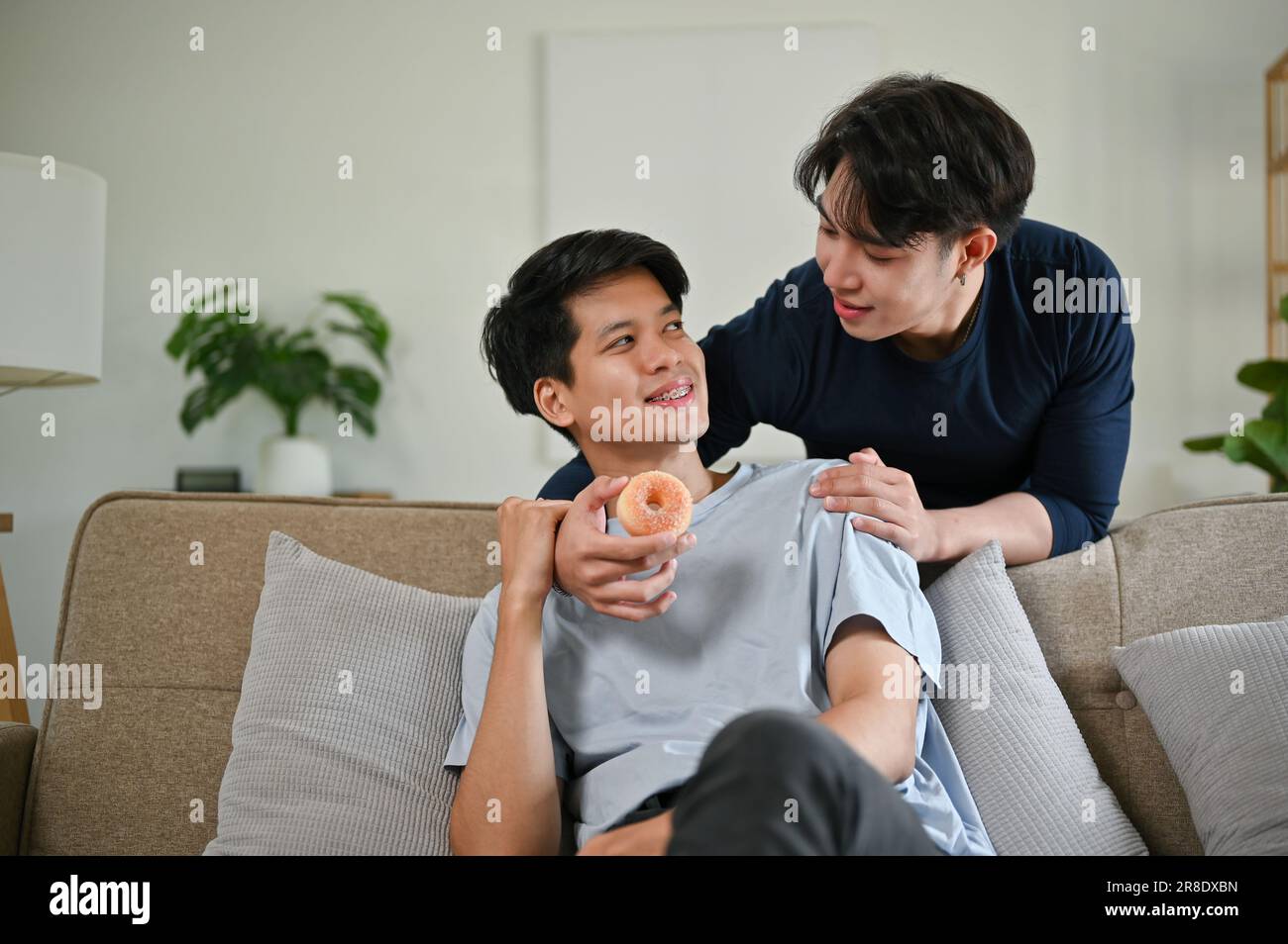 Un bel giovane gay asiatico, felice e simpatico, nutre una ciambella al suo ragazzo mentre si rilassa su un divano nel soggiorno insieme. LGBTQ+, stile di vita, c Foto Stock