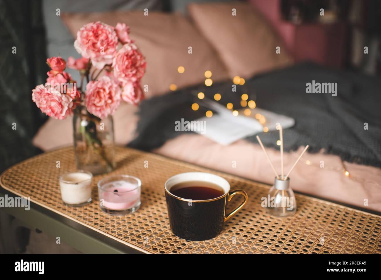 Tazza di tè con candela profumata e bouquet di fiori di rosa sul tavolo di rattan sul letto con illuminazione closeup. Atmosfera accogliente e igge. Foto Stock