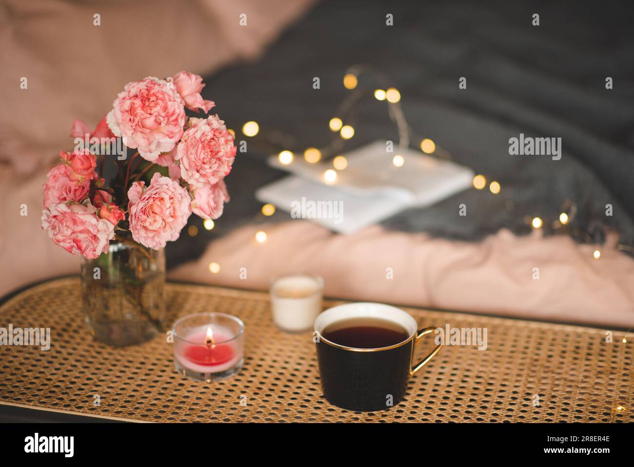 Tazza di tè con candela profumata e bouquet di fiori di rosa sul tavolo di rattan sul letto con illuminazione closeup. Atmosfera accogliente e igge. Foto Stock