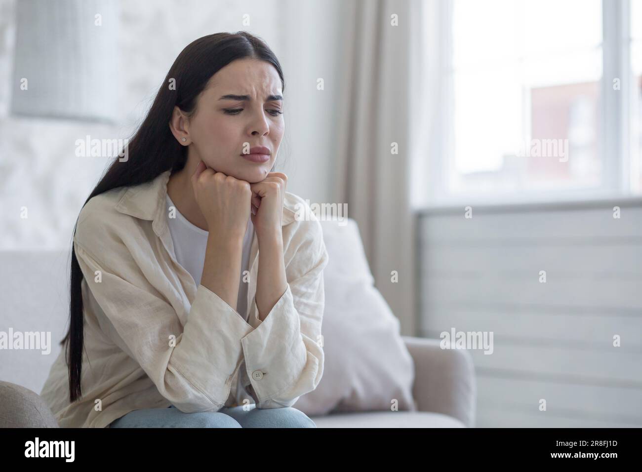 Giovane donna sconvolta seduta sul divano a casa, occhi chiusi, mani ripiegate accanto al viso. Soffre di depressione, piange, pensa a risolvere i problemi. Foto Stock