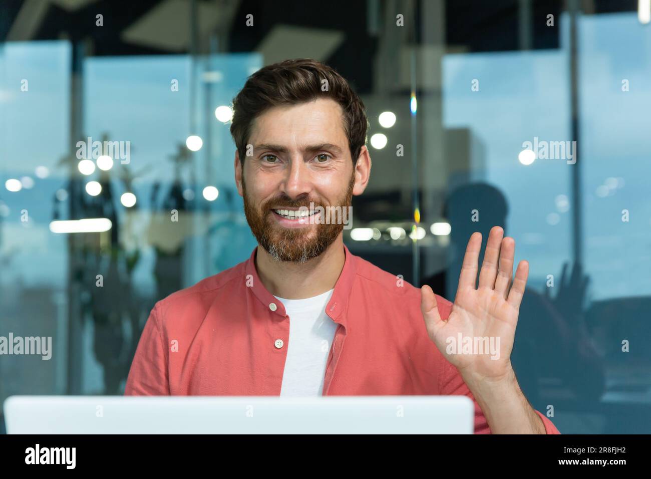 Ritratto di un giovane programmatore maschio in una camicia rossa che lavora in ufficio a un computer portatile. Guarda la telecamera con un sorriso, gli fa una mano. Primo piano. Foto Stock