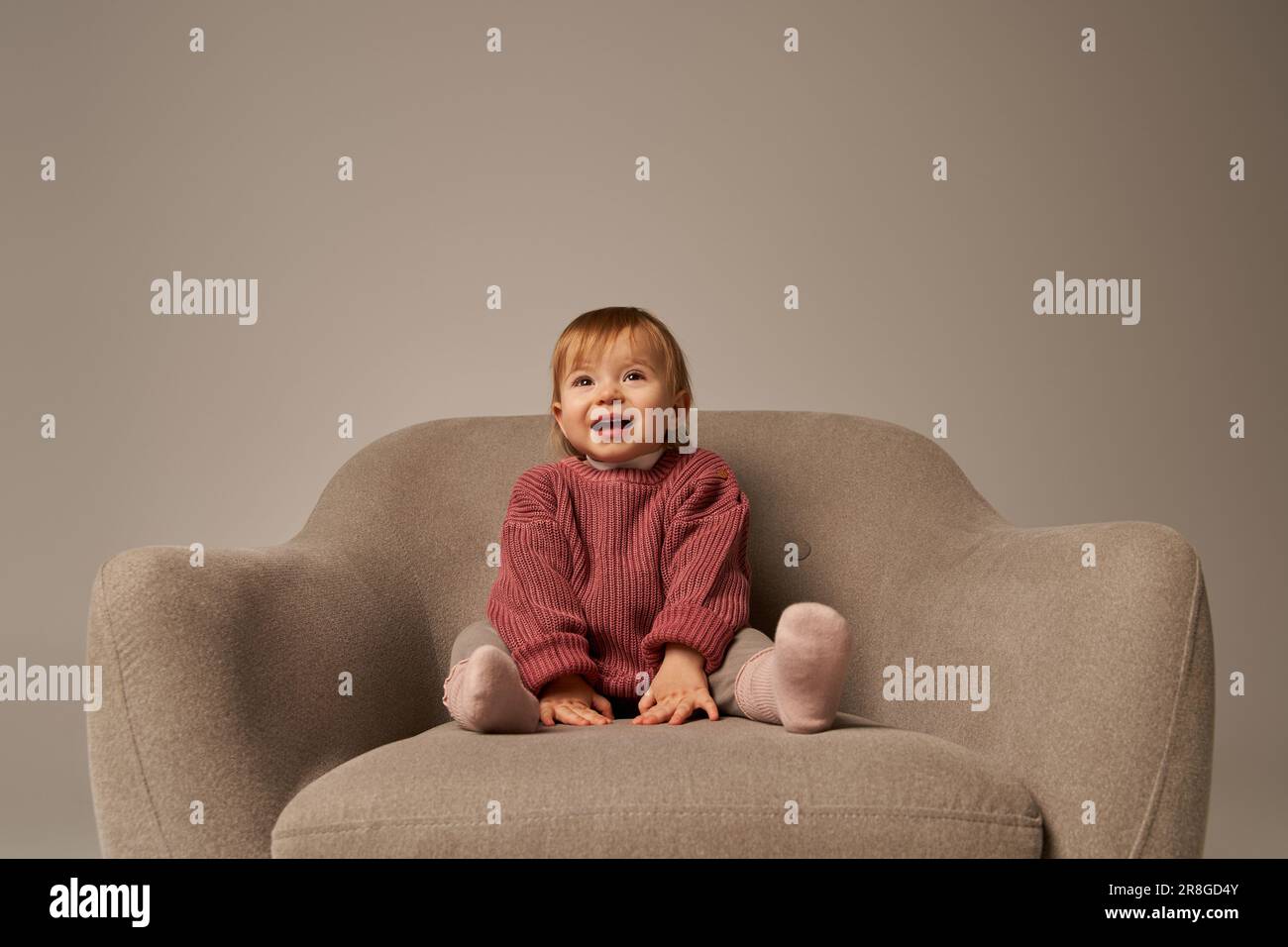 ragazza cute del bambino, toddler in abbigliamento casual seduto su comoda poltrona su sfondo grigio in studio, emozione, felicità, gioia, innocenza, piccolo chil Foto Stock