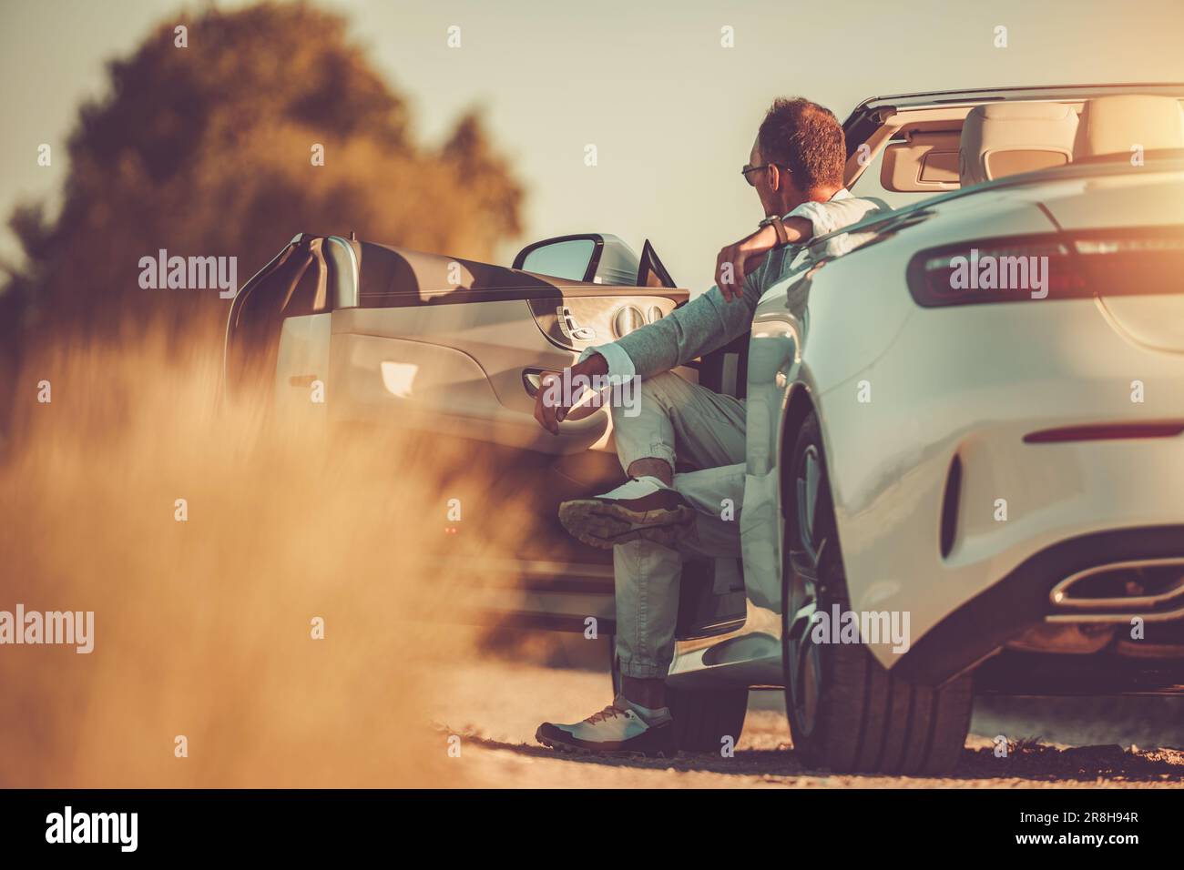 Uomo di mezza età di successo e ricco seduto nel suo moderno bianco convertibile godendo colorato tramonto. Vita felice. Foto Stock