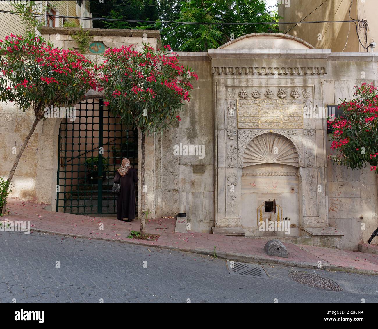 Donna che indossa un hijab in una strada con una fontana e accesso recintato a una proprietà, più alberi con fiori rossi. In Turchia Foto Stock