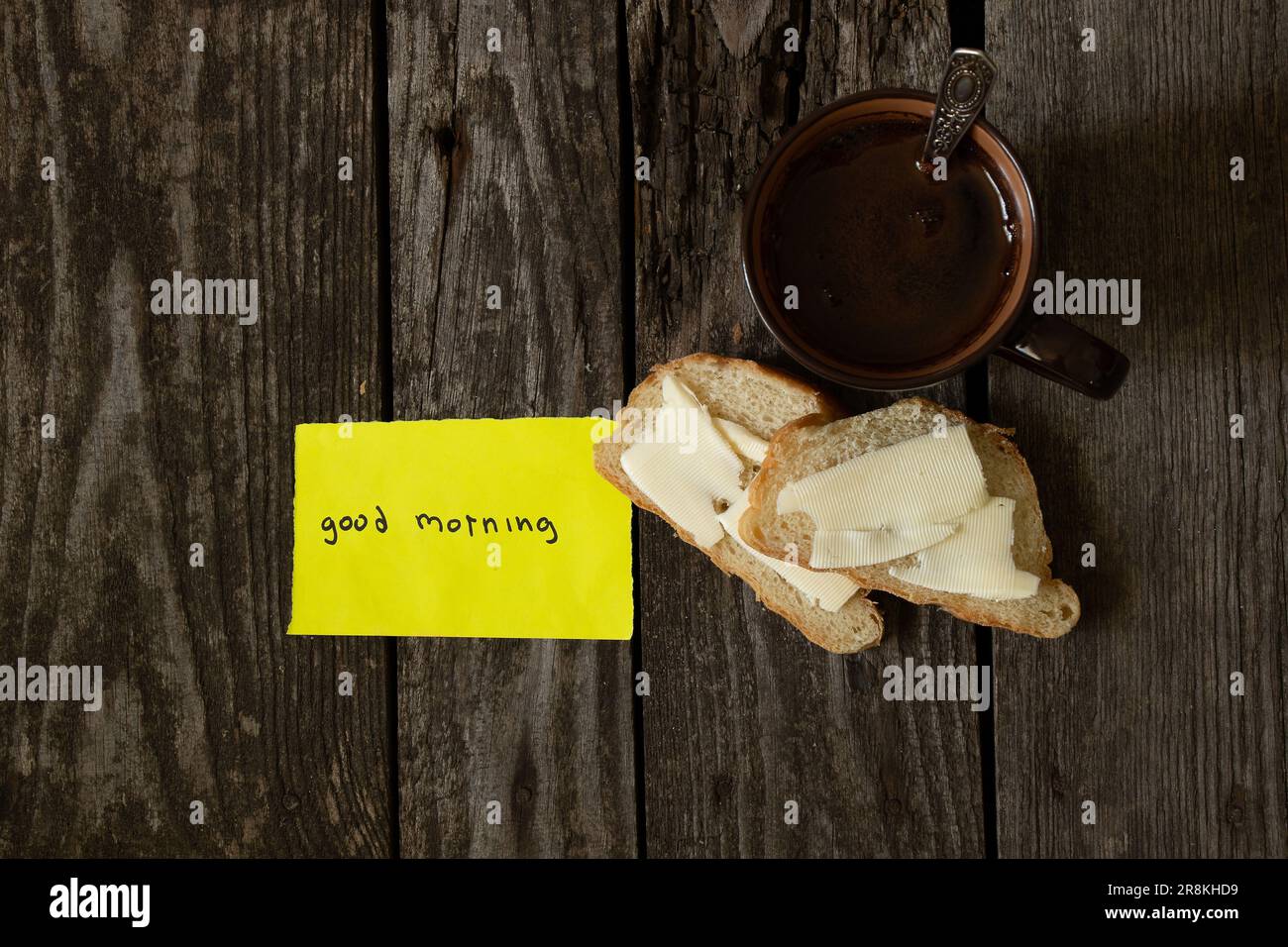 Caffè nero caldo e pane bianco e burro su un tavolo di legno e le parole in inglese Buongiorno su carta gialla Foto Stock