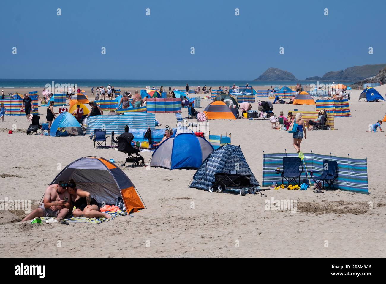 I turisti potranno godersi il sole sulla spiaggia di Peranporth, Cornovaglia, Inghilterra. I frangivento multicolore e gli ombrelli proteggono dal calore. Foto Stock