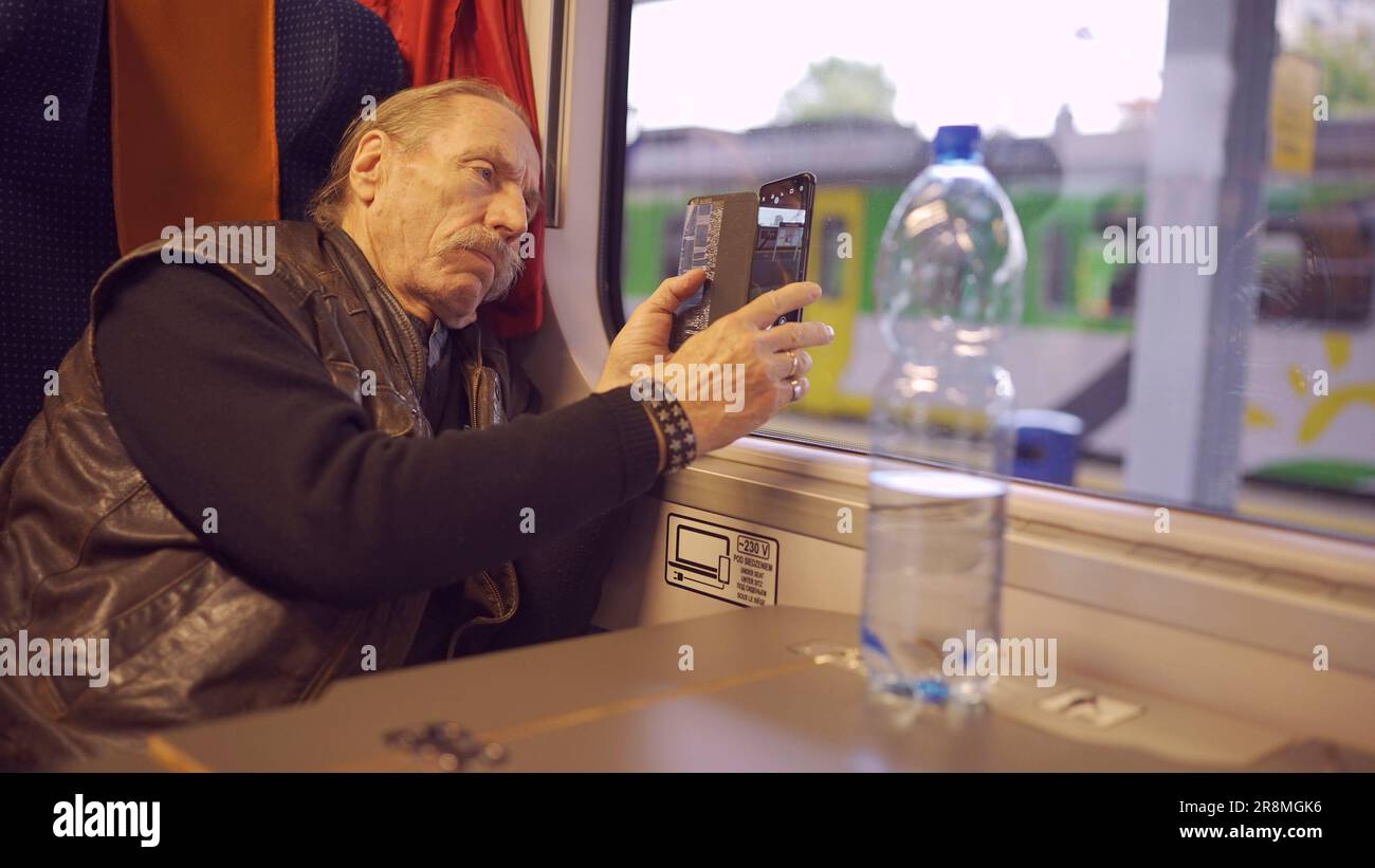 Uomo anziano che viaggia in treno e scatta foto del paesaggio attraverso la finestra utilizzando uno smartphone Foto Stock