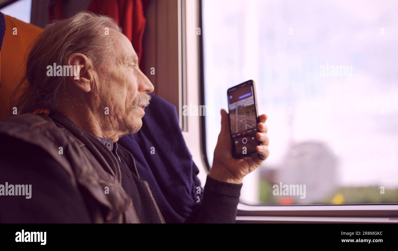Uomo anziano che viaggia in treno e scatta foto del paesaggio attraverso la finestra utilizzando uno smartphone Foto Stock