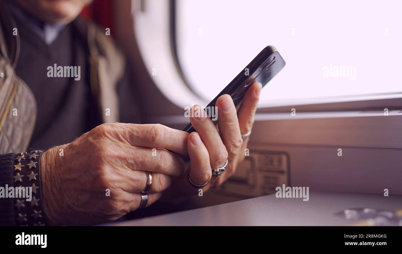 Primo piano delle mani di un anziano seduto in un carraageriage ferroviario e utilizzando uno smartphone Foto Stock