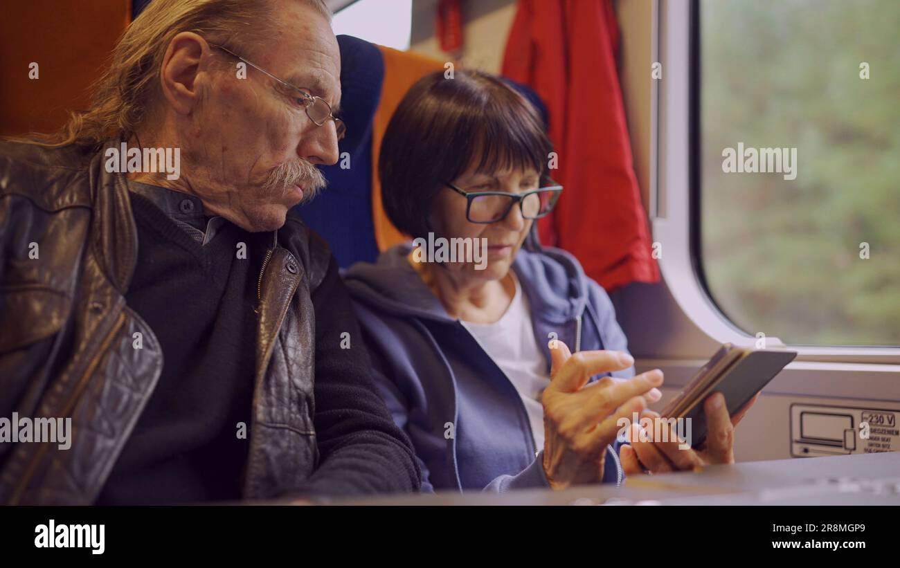 La coppia anziana viaggia in treno, la signora tiene in mano un telefono cellulare, entrambi guardano lo smartphone e parlano tra loro Foto Stock