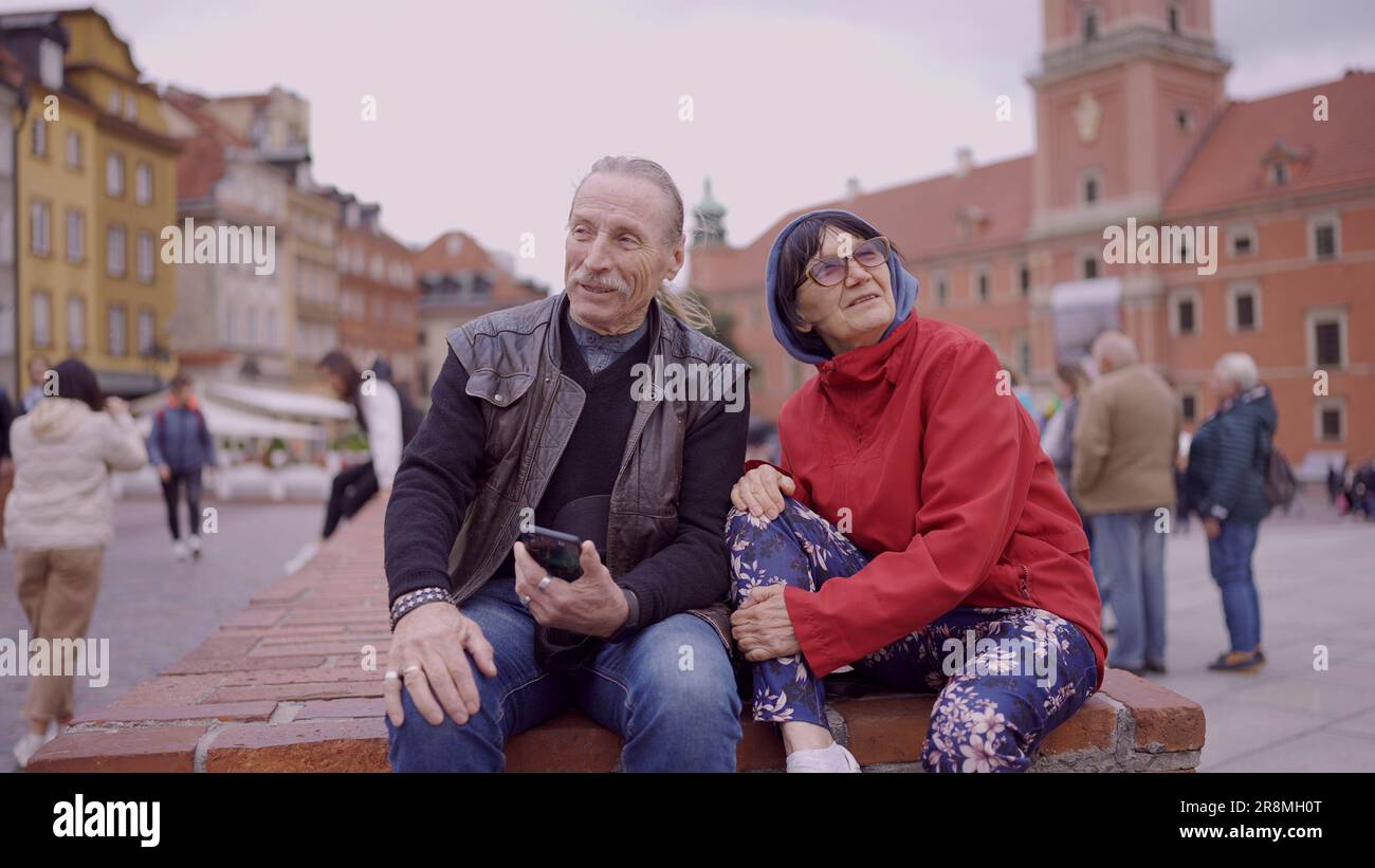Una coppia di turisti anziani è seduta e parla nel centro storico di una vecchia città europea, senior ha uno smartphone in mano. Piazza del Palazzo, Foto Stock