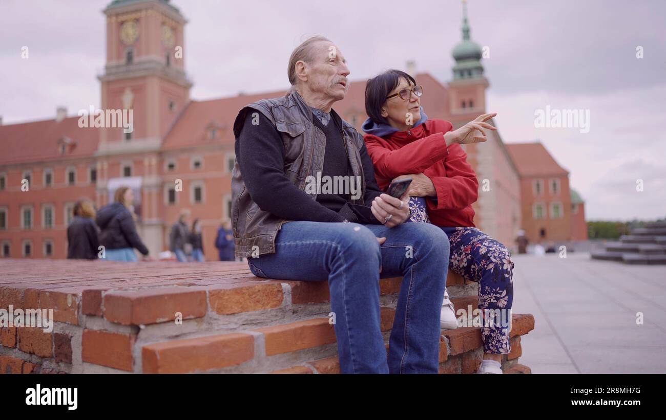 Una coppia di turisti anziani è seduta e parla nel centro storico di una vecchia città europea, senior ha uno smartphone in mano. Piazza del Palazzo, Foto Stock
