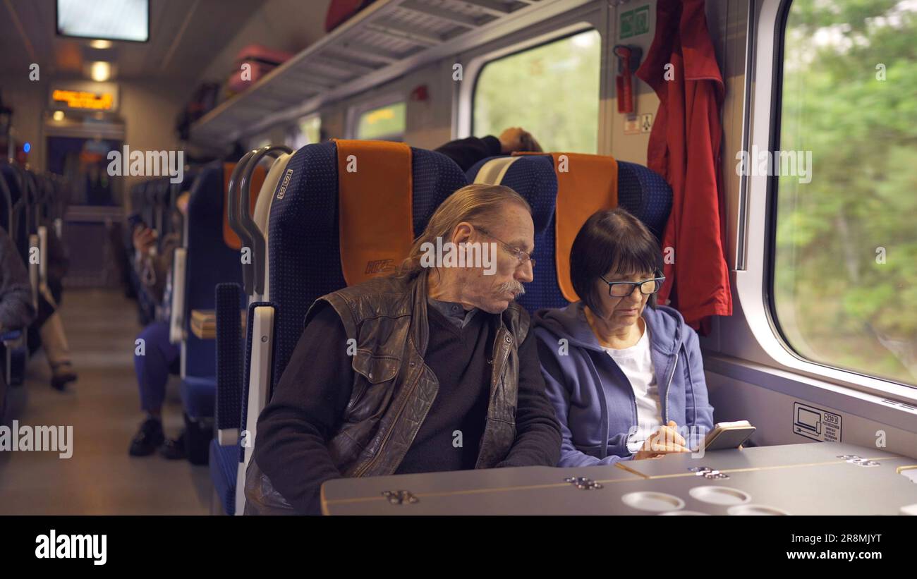La coppia anziana viaggia in treno, la signora tiene in mano un telefono cellulare, entrambi guardano lo smartphone e parlano tra loro Foto Stock