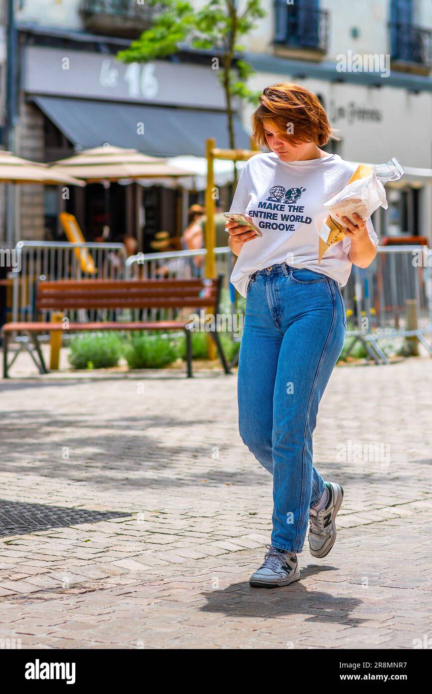 Giovane donna in T-shirt e jeans in possesso di pane, dolci e guardare il telefono cellulare mentre si cammina nel centro della città - Tours, Indre-et-Loire (37), Francia. Foto Stock