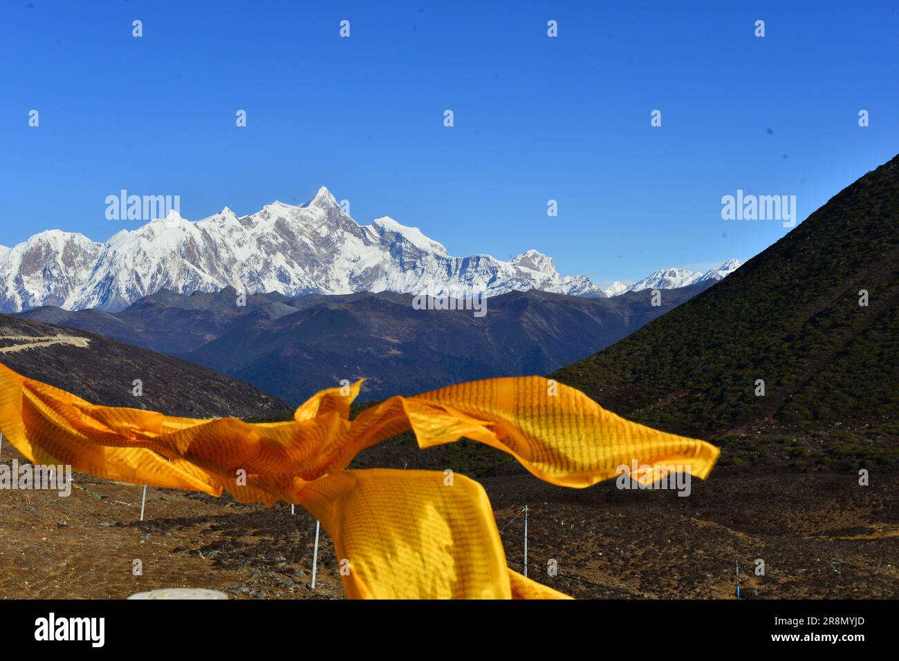 Le bandiere di preghiera dorate sventolano nel vento sullo sfondo di maestose montagne innevate e di un cielo blu profondo, che simboleggiano la spiritualità e la natura» Foto Stock