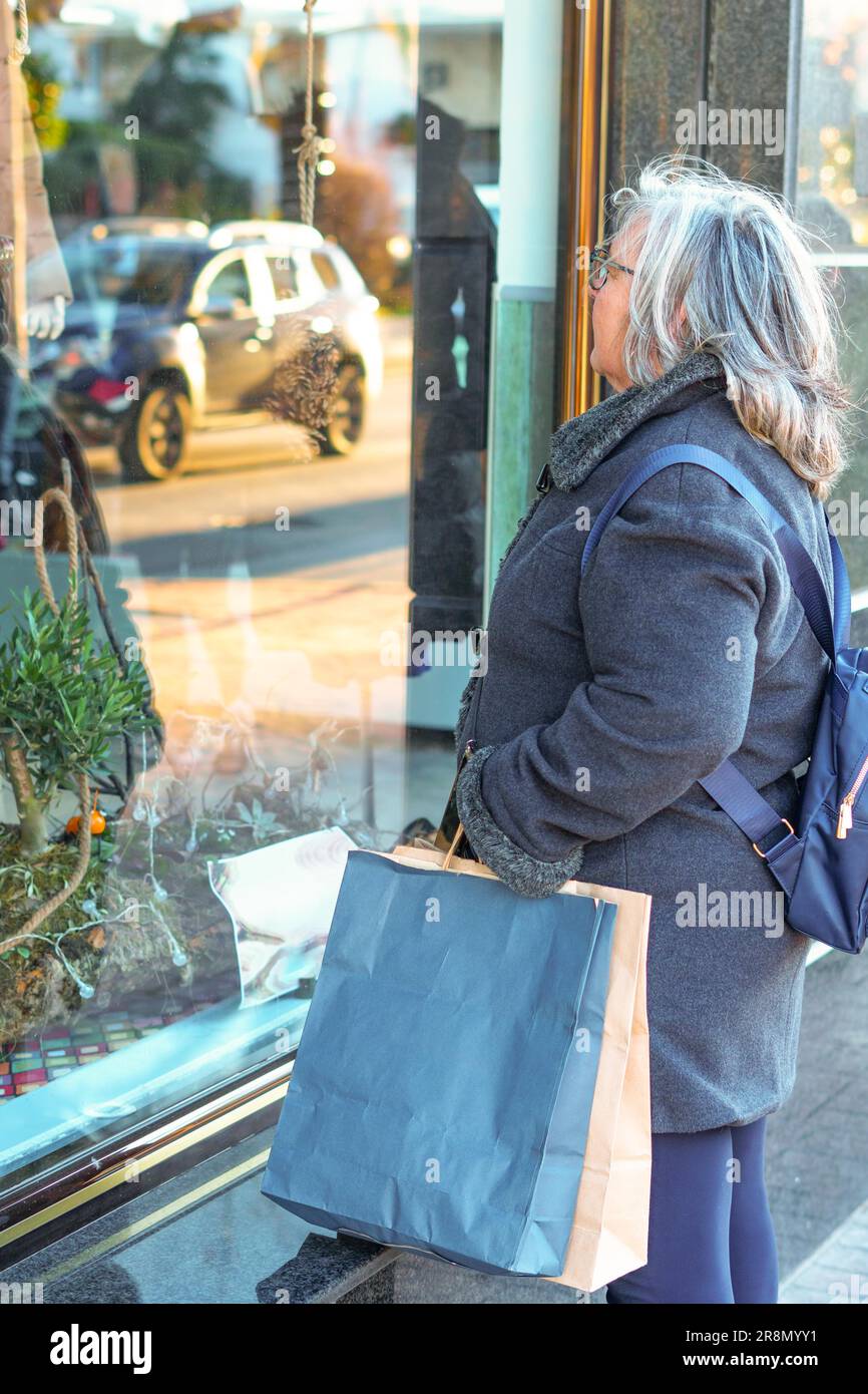 Ronda, malaga, spagna donna più anziana in capelli bianchi e cappotto con borse per la spesa in una vetrina Foto Stock