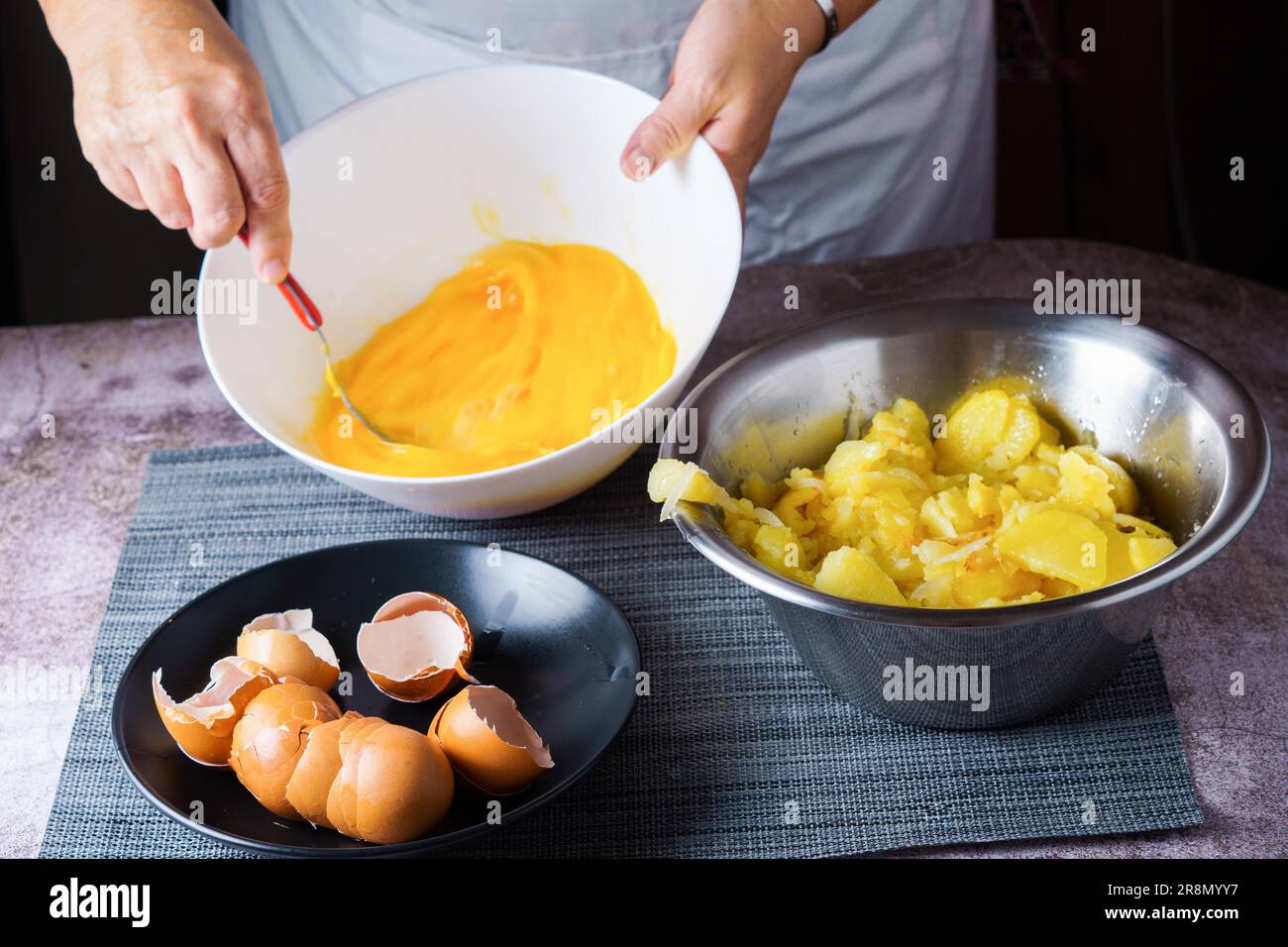 Donna in grembiule bianco che sbatte un uovo in una ciotola con un piatto di gusci d'uovo e una ciotola di patate per fare una frittata Foto Stock