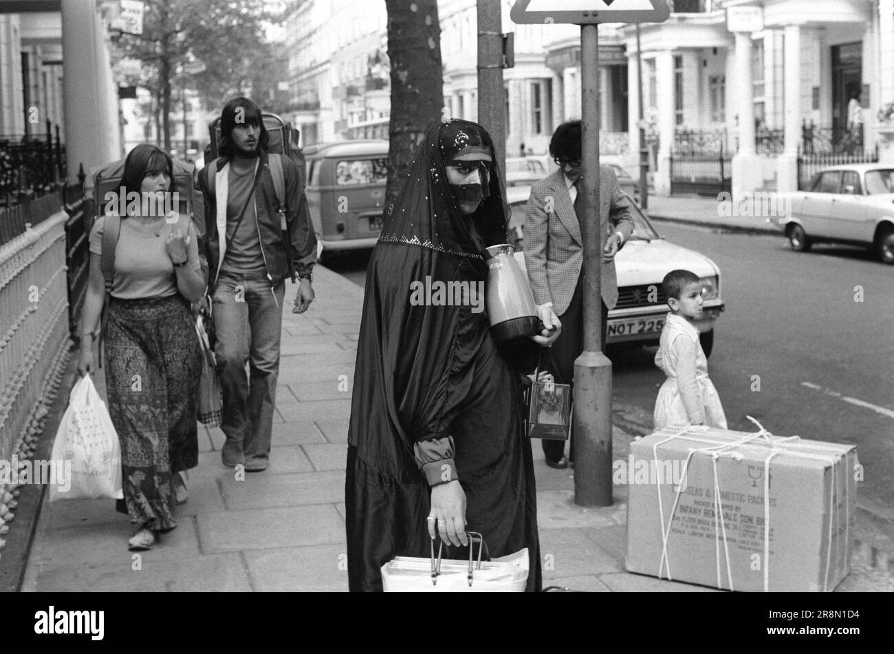Arab Family Earls Court, Londra sta per partire con cose acquistate mentre nel Regno Unito 1970s. La gente del Medio Oriente è venuta in Gran Bretagna per assistenza sanitaria sovvenzionata nelle cliniche di Harley Street. Sono stati principalmente in alberghi economici a Earls Court. Due backpackers vagano oltre una donna araba che indossa una maschera Battoulah che di solito indica che è sposata. Earls Court, Londra, Inghilterra circa 1977 70s Regno Unito HOMER SYKES Foto Stock