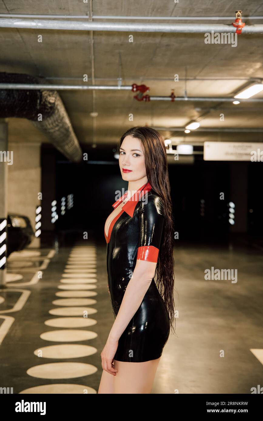 Una donna bruna che indossa un abito in lattice nero e rosso si trova in un garage sotterraneo Foto Stock