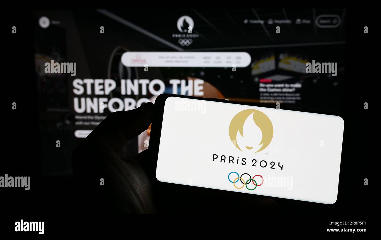 Persona che possiede un telefono cellulare con il logo delle Olimpiadi estive di Parigi del 2024 sullo schermo davanti alla pagina Web. Concentrarsi sul display del telefono. Foto Stock