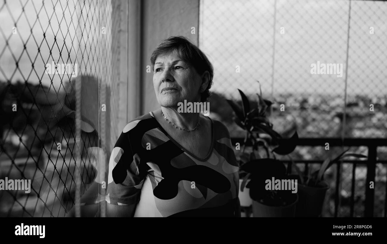Triste donna anziana depressa che si sente sola a casa in piedi accanto alla finestra guardando la vista della città in una profonda introspezione. Anziana signora in pensione in monocromatico Foto Stock