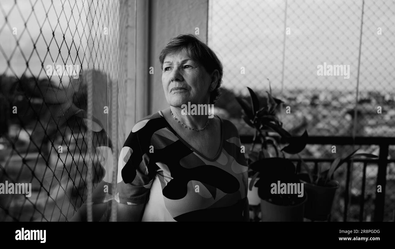 Triste donna anziana depressa che si sente sola a casa in piedi accanto alla finestra guardando la vista della città in una profonda introspezione. Anziana signora in pensione in monocromatico Foto Stock