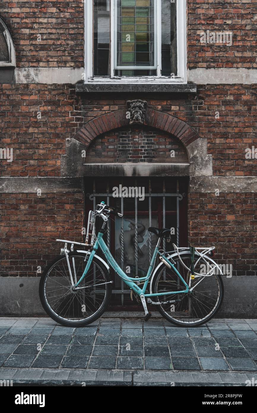 In uno stretto vicolo di Brugge, in Belgio, una solitaria bicicletta blu si appoggia contro un muro di mattoni, pazientemente in attesa del ritorno del pilota. Foto Stock