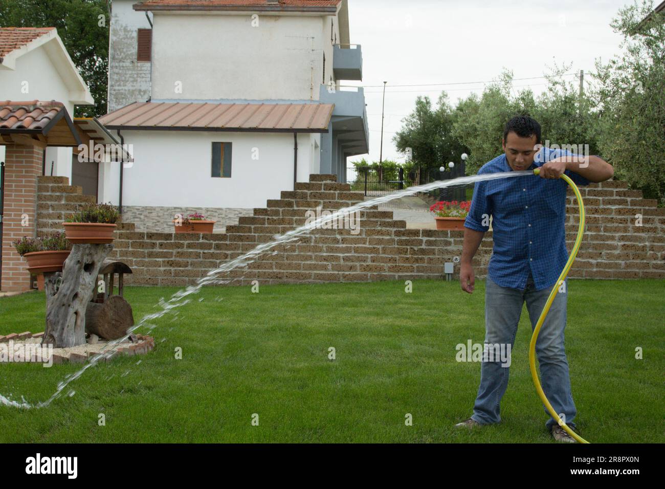 Immagine di un uomo nel suo giardino di casa che beve acqua da un tubo per prato. Riferimento ai rifiuti idrici. Foto Stock