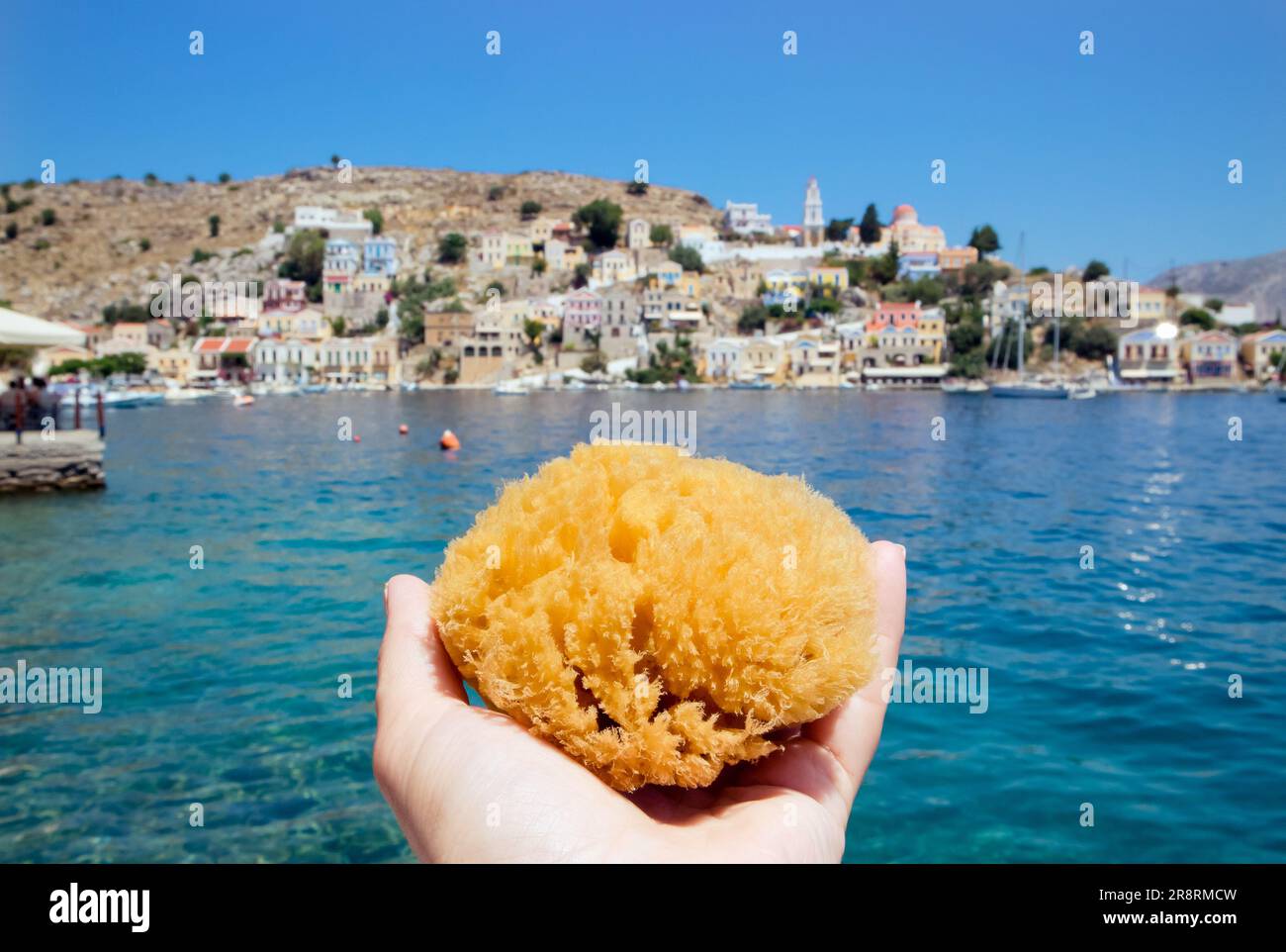 Turista in possesso di una spugna marina greca dell'isola di Symi, con la città di Symi sullo sfondo nelle soleggiate giornate estive. Symi è popolare per la sua spugna marina. Foto Stock