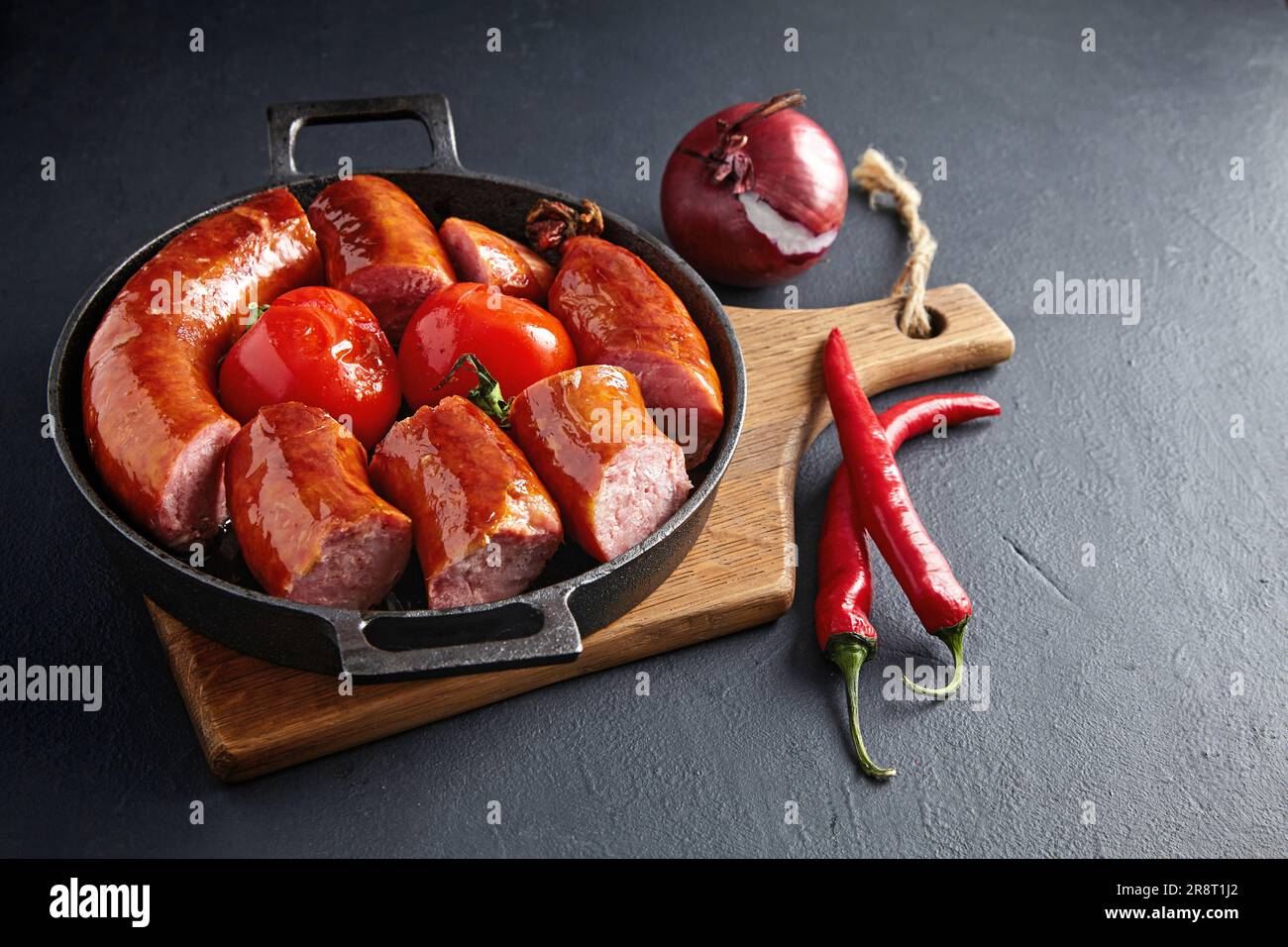 salsiccia di maiale fritta a fette in una padella di ghisa con pomodori su un tagliere di legno su un tavolo di pietra nera Foto Stock