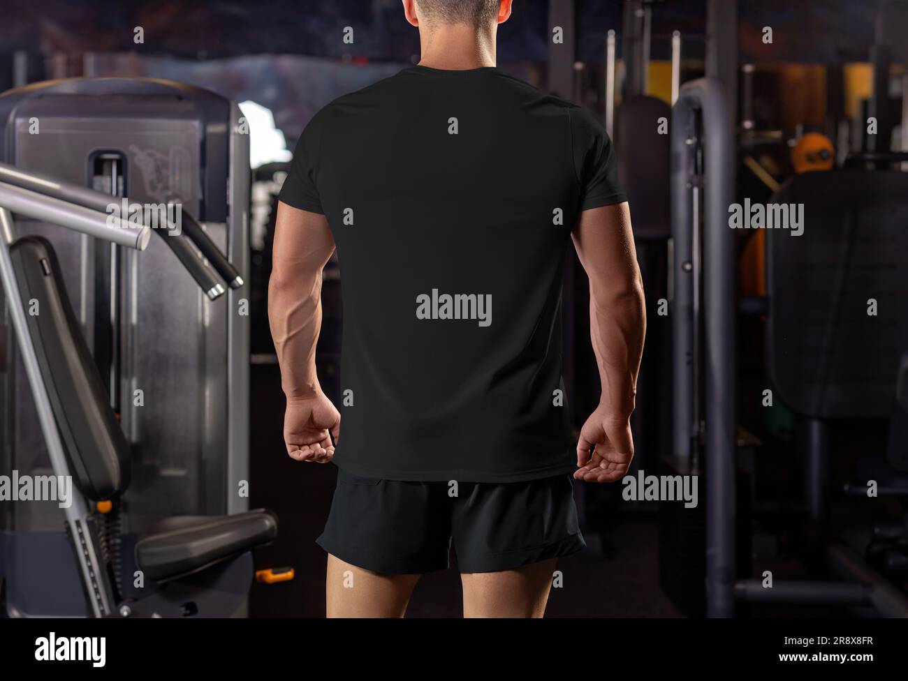 Mockup di una t-shirt nera su un ragazzo muscoloso sullo sfondo dell'attrezzatura in palestra, vista posteriore. Modello di abbigliamento sportivo per l'allenamento. ph. Prodotto Foto Stock