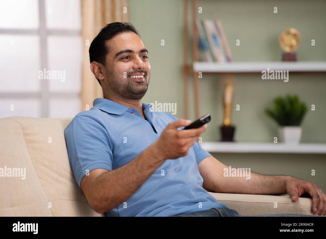 Rilassato uomo indiano felice che guarda le partite sportive dal vivo in tv o in televisione mentre si siede sul divano a casa - concetto di intrattenimento, attività ricreative e.. Foto Stock