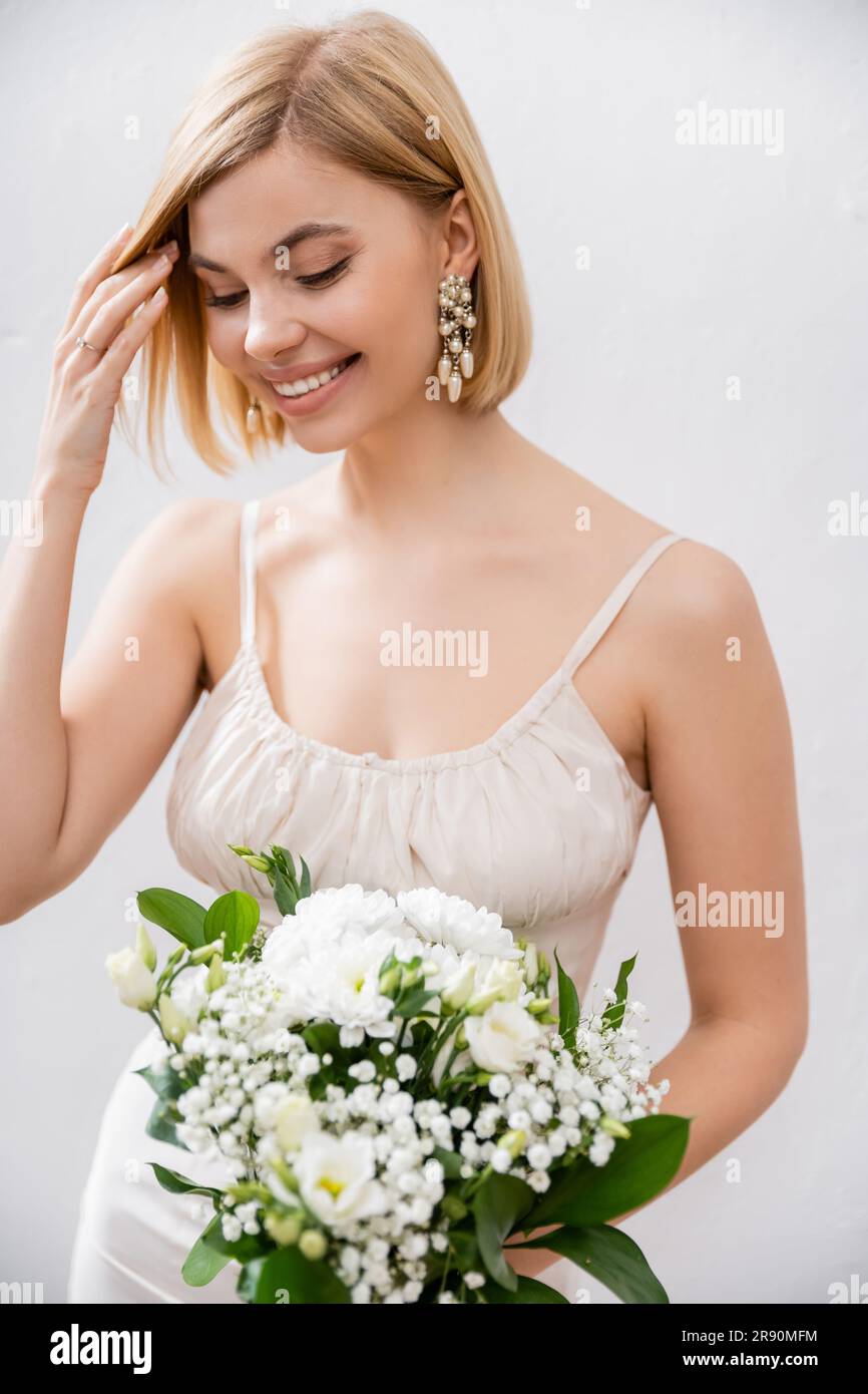 sposa allegra e bionda in abito da sposa con bouquet su sfondo grigio, fiori bianchi, accessori da sposa, felicità, occasione speciale, beaut Foto Stock