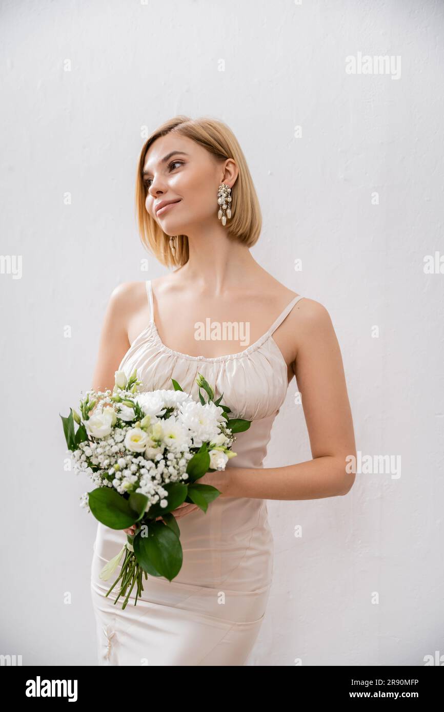 sposa bella e bionda in abito da sposa con bouquet su sfondo grigio, fiori bianchi, accessori da sposa, felicità, occasione speciale, bea Foto Stock