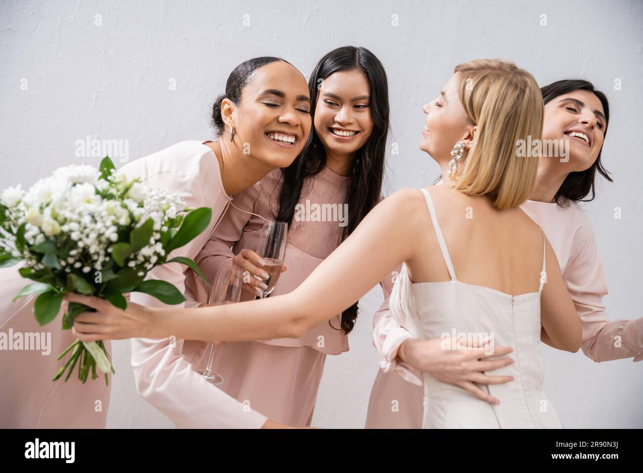 preparativi per matrimoni, sposa felice con bouquet che abbraccia allegri damigelle multiculturali con champagne su sfondo grigio, vestibilità, damigella d'onore Foto Stock