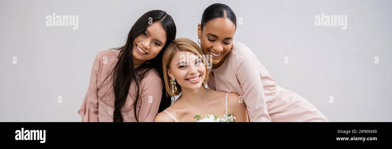 fotografia di matrimonio, diversità culturale, tre donne, felice sposa con bouquet e le sue damigelle interrazziali sedute su poltrona su sfondo grigio, Foto Stock