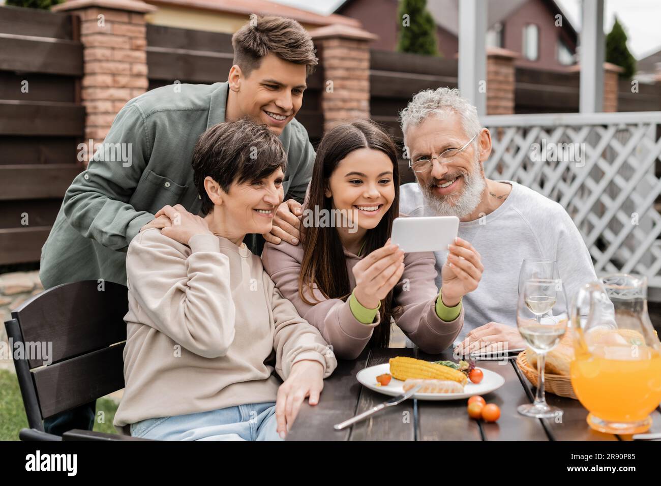 Adolescente sorridente che usa lo smartphone vicino a genitori di mezza età e fratello durante la festa al barbecue con cibo estivo e festa dei genitori nel cortile Foto Stock