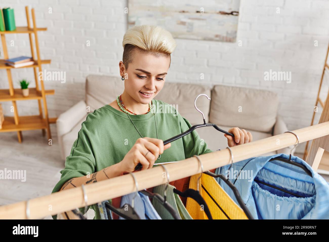 donna tatuata con un sorriso felice e un appendiabiti alla moda vicino al rack con abbigliamento durante il processo di decluttering in salotto, fa sostenibile Foto Stock