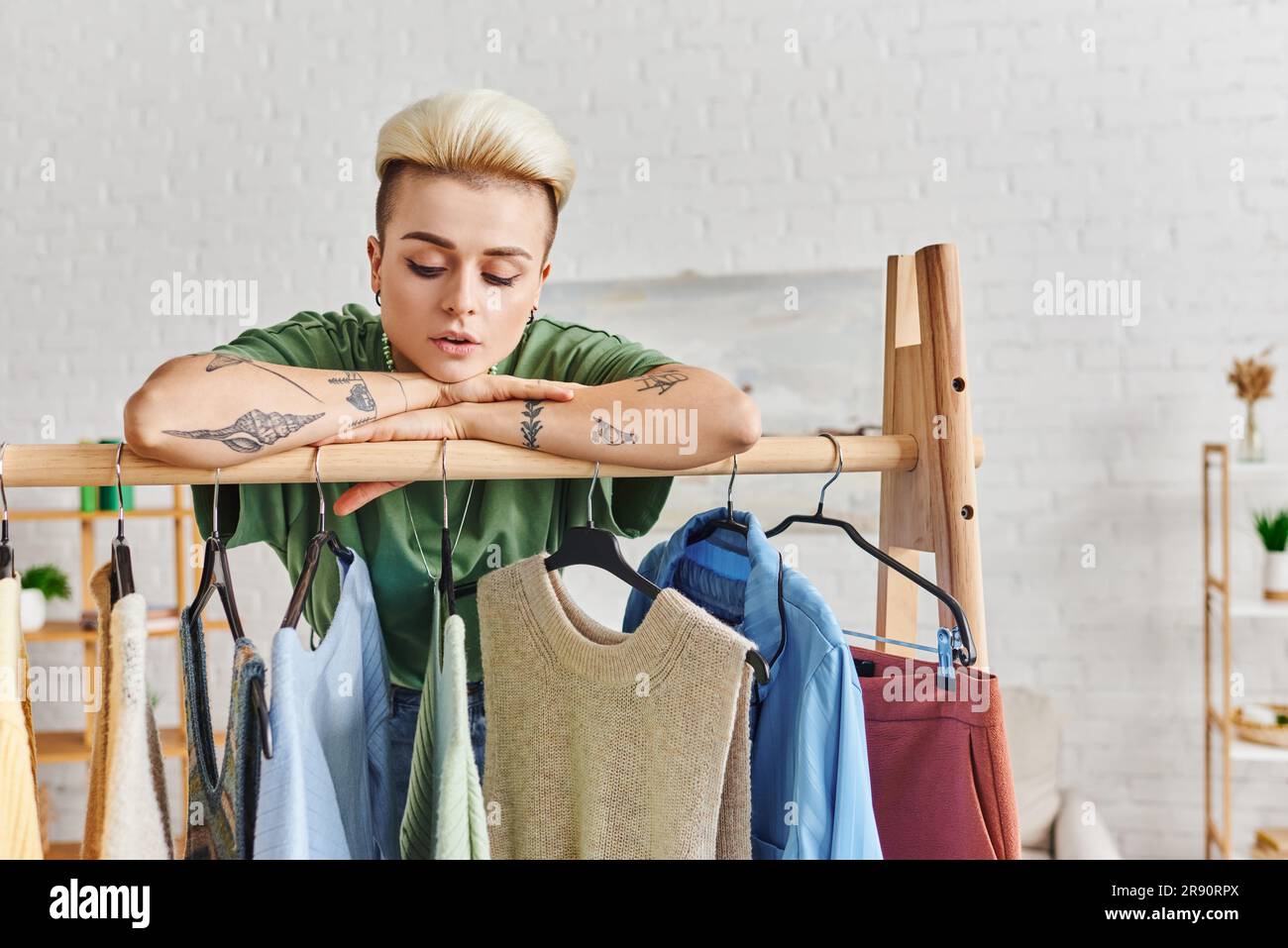 guardaroba, donna tatuata da sogno con acconciature alla moda appoggiata su un rack con abiti casual alla moda su grucce, moda sostenibile Foto Stock