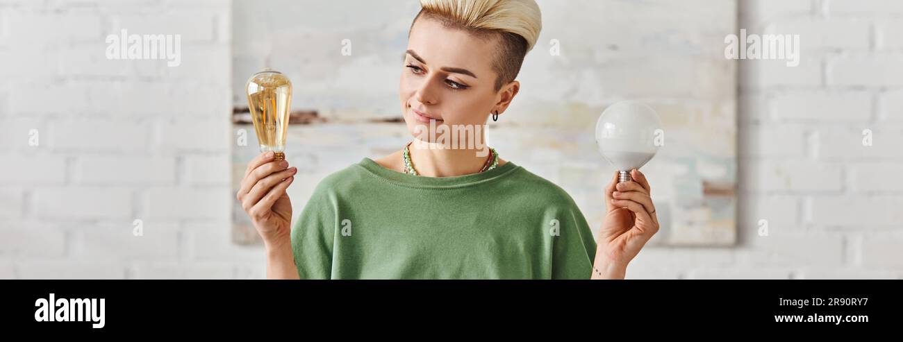 donna giovane e sorridente con un'acconciatura alla moda che confronta le lampadine a risparmio energetico mentre si trova a casa, scelte ecocompatibili e uno stile di vita sostenibile Foto Stock
