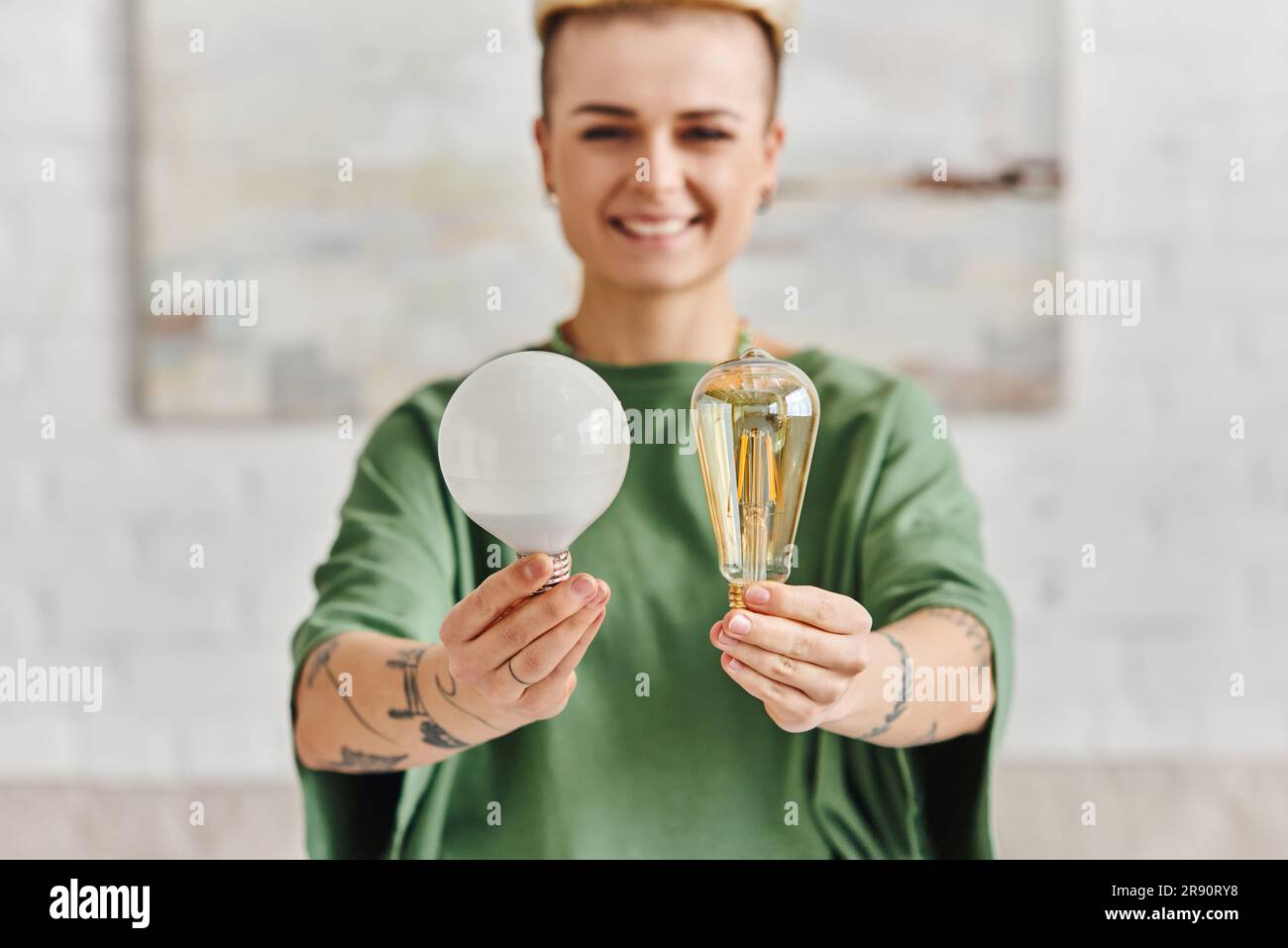 donna allegra, giovane e tatuata in abiti casual che mostra lampadine a risparmio energetico e guarda la fotocamera, rispettosa dell'ambiente, sostenibile Foto Stock