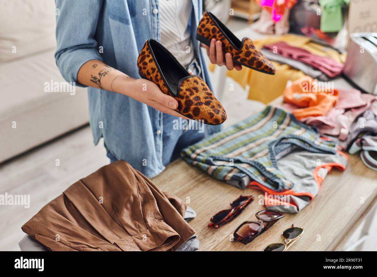 consumo etico, scambio, vista ritagliata di una giovane donna tatuata che tiene eleganti scarpe con stampe animali vicino a occhiali da sole e indumenti sul tavolo, sustaina Foto Stock